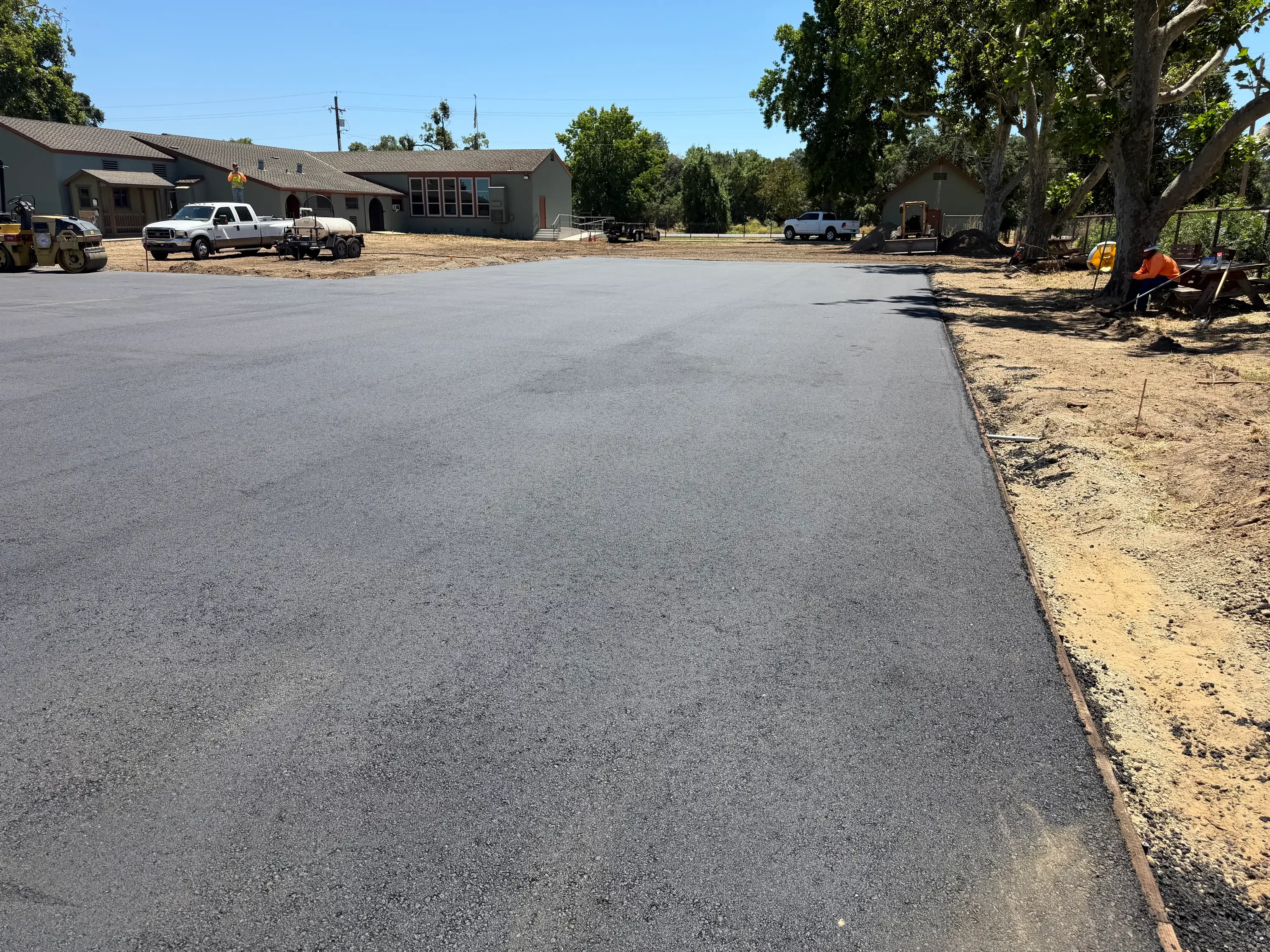 Newly paved asphalt driveway with commercial buildings and mature trees in background